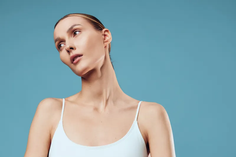 Young woman with natural skin tone posing against blue background, wearing white tank top, minimal makeup, soft lighting, contemplative expression, clean and simple style