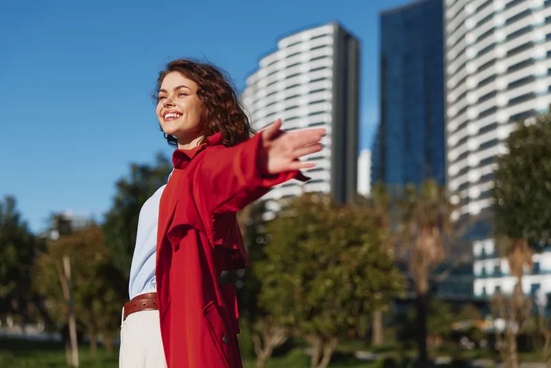 Joyful woman with open arms stands outdoors in a redcoat against a sunlit cityscape, embodying freedom and upbeat energy as glass towers glow under a clear blue sky today.