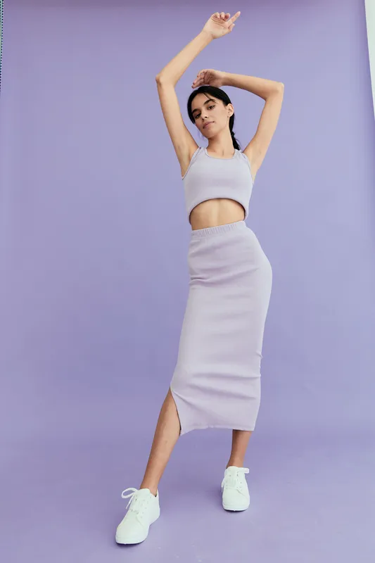 young woman in casual lavender outfit posing confidently against purple background, wearing sleeveless crop top and midi skirt with white sneakers, minimalistic style