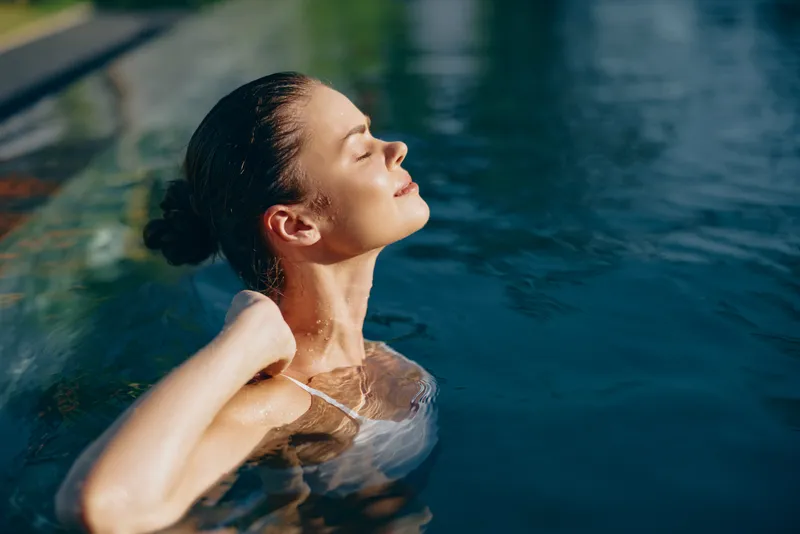 Young woman with wet hair relaxes in a swimming pool with closed eyes, enjoying a peaceful moment of calm and mindfulness in warm natural light.