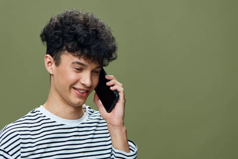 Young man talking on smartphone, smiling with curly hair, wearing striped shirt, isolated on green background, casual communication moment.