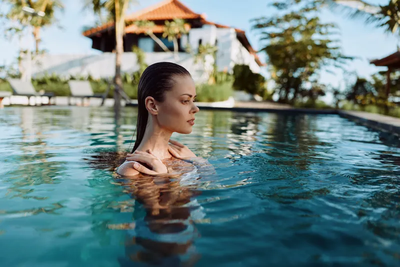 Young woman with dark hair floats in a tranquil pool, gazing aside as a tropical resort backdrop frames a serene moment