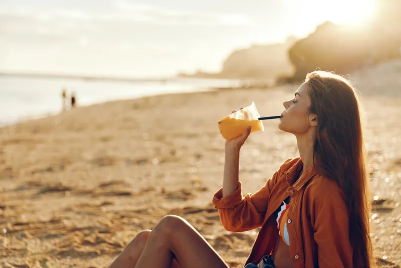 Woman drinking water from bottle at sunset on beach outdoors landscape recreation lifestyle, summer travel seaside coast ocean waves, sunlight golden evening, relaxed female adult in jacket sitting