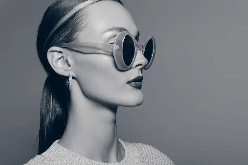 Profile of a young white woman with pale skin wearing round sunglasses and earrings in monochrome studio portrait.
