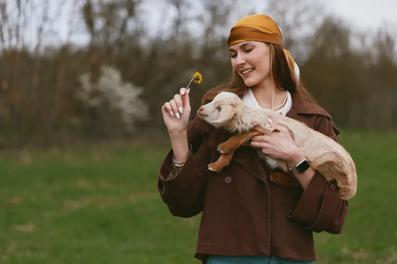 Cheerful woman outdoors, holding a playful goat kid, smiling at a flower, sunlit meadow scene, casual jacket, relaxed mood, natural colors, countryside lifestyle moment