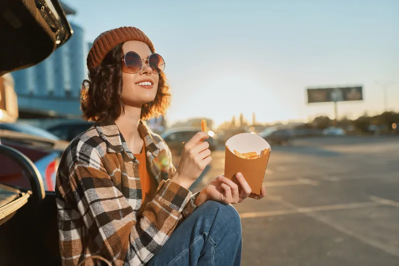 Woman eating fries by car trunk in parking at sunset, sunglasses and plaid jacket, casual smile and urban vibe. Authenticity, candid lifestyle, golden hour glow, mindful living.