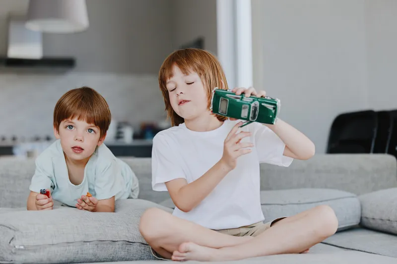 Two happy boys playing at home, one holding a camera while the other plays with colorful toys. Natural light creates a warm, playful atmosphere.
