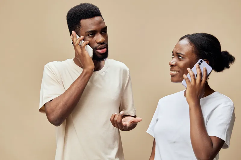 Young Black couple talking on smartphones, expressing emotions, stylish casual clothing, warm beige background, modern communication concept
