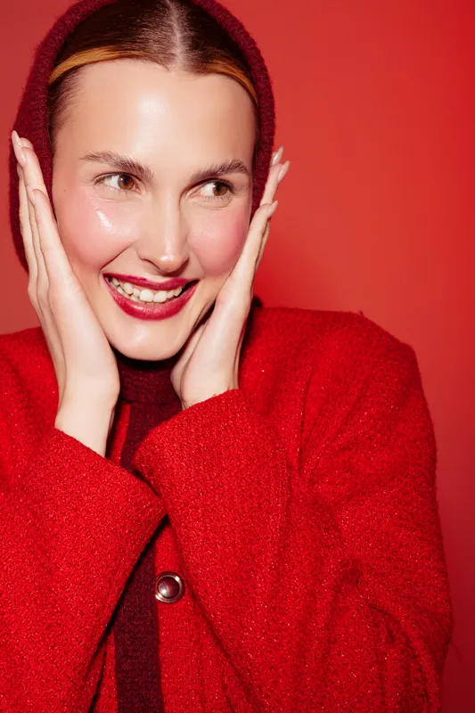 Smiling woman in red wool coat and hood touches face with hands, looking sideways. Bright red background and warm clothing create cozy winter fashion concept with joyful mood and natural beauty.