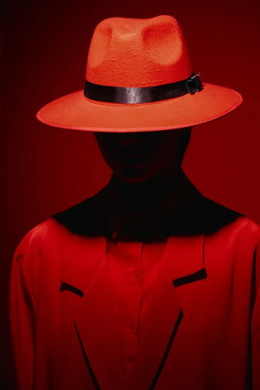 Mysterious woman in red coat wears red fedora hat partially hiding her face against red background. Creative beauty and fashion concept with dramatic lighting.