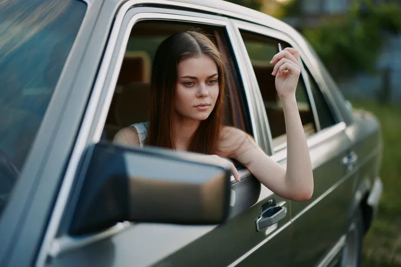 Thoughtful young woman with long hair sitting in a car, holding a cigarette against a blurred green background, embodying a sense of nostalgia and contemplation.