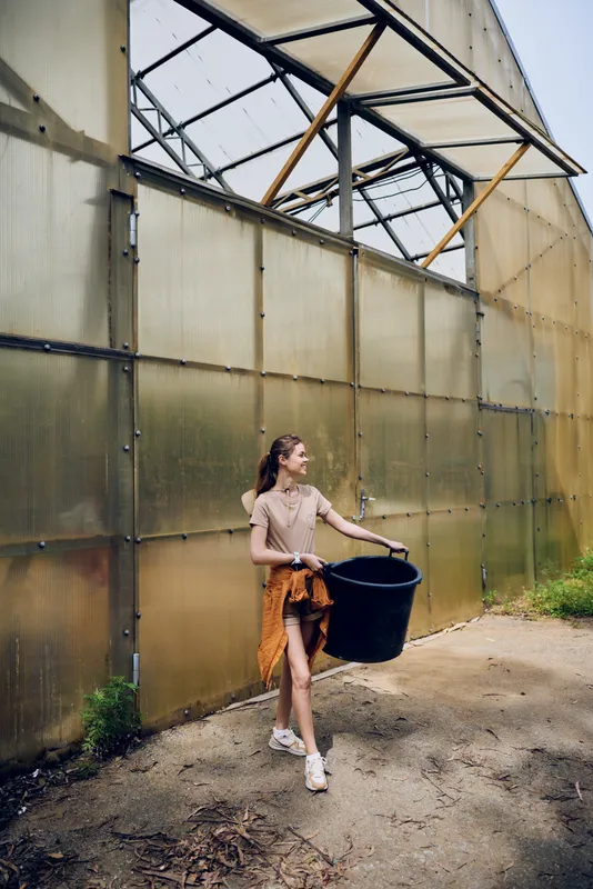 Woman girl in greenhouse carrying a black bucket for gardening on a small farm, casual summer outfit and sneakers, young female walking along polycarbonate wall with plants nearby.