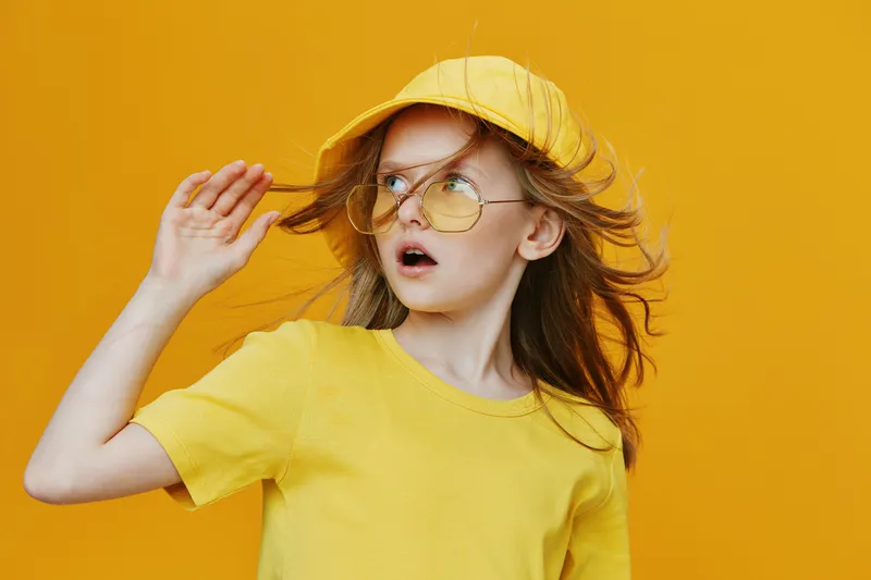 Young girl wearing a yellow cap and sunglasses, surprised expression, with flowing hair against a bright yellow background, perfect for summer fashion or kids wear