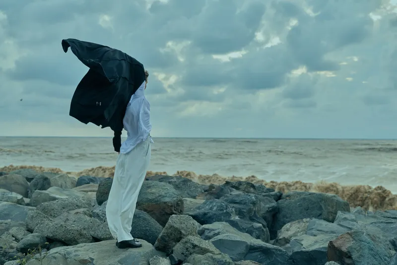 Windswept figure on a rocky coastline, coat blown over the head, a moody outdoor scene by the sea with dramatic overcast weather and isolated silhouette