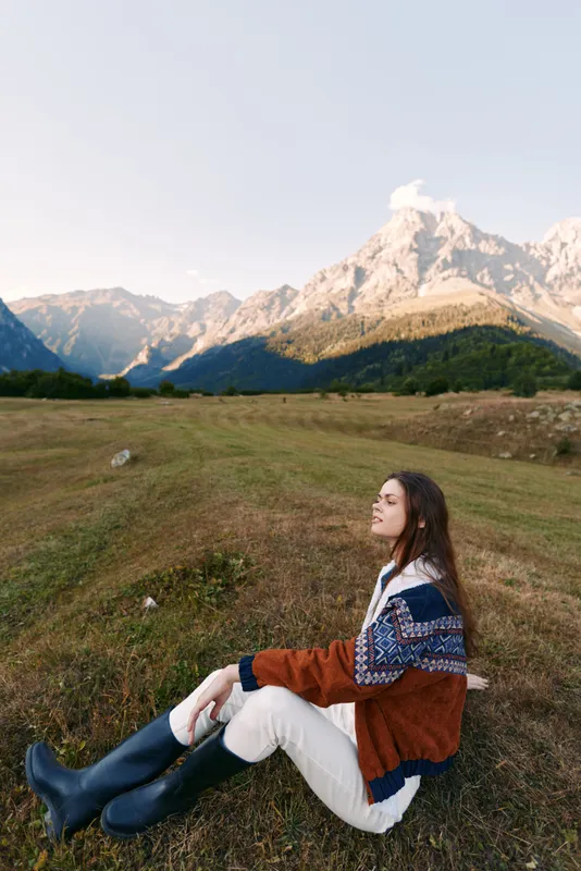 Woman sitting in a meadow wearing a patterned sweater and rubber boots, enjoying mountains and open field at golden hour, serene outdoor portrait and travel moment in nature