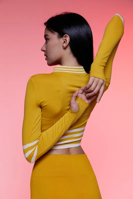 Woman sport fitness wearing yellow activewear stretching arms with focused expression on pink background, showcasing flexibility and strength for healthy lifestyle.