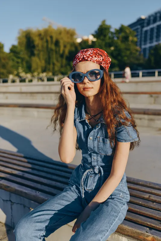 Lifestyle woman wearing modern boho-western denim street style posing outdoors with oversized sunglasses and red bandana under warm sunlight, film color effect applied
