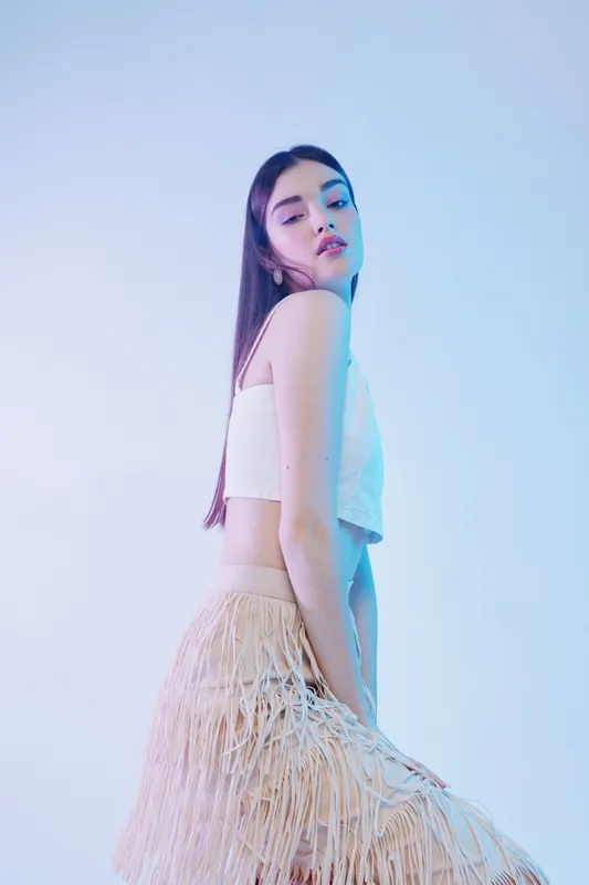 Young woman in a white crop top and fringed skirt poses confidently in soft studio lighting. Concept of beauty, fashion, and modern style with a minimalistic background.