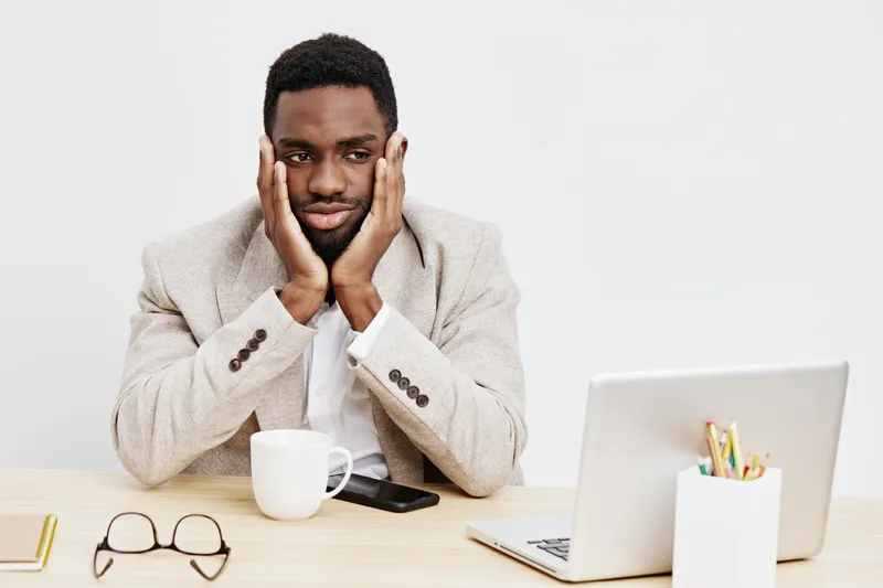 Frustrated young man with dark skin sits at desk with laptop, holding head in hands in discomfort. Office workspace with white background, coffee mug, glasses, and stationery, concept of stress, work 