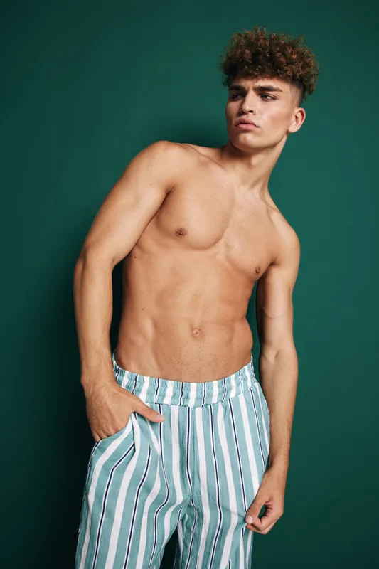 Young man with curly hair posing shirtless against green background wearing striped pants with hand in pocket looking away with confident expression and relaxed posture.