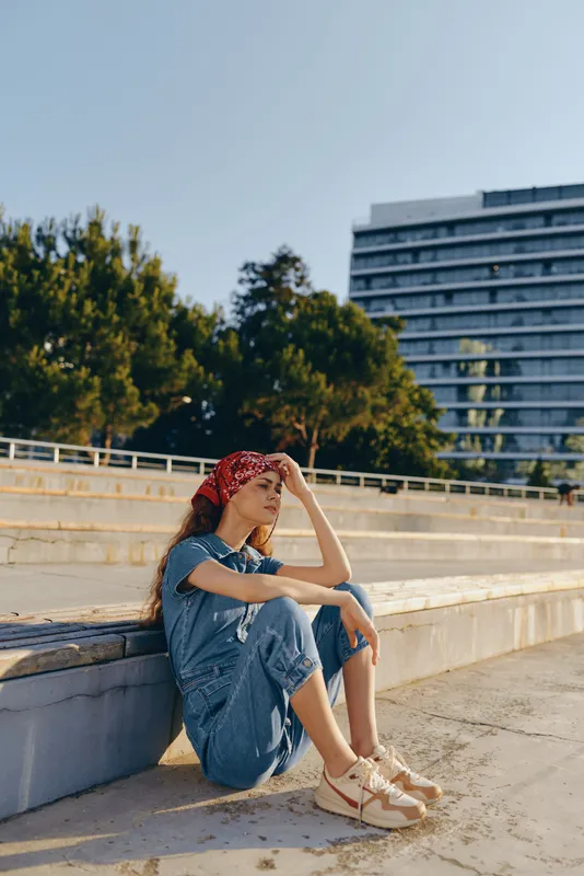 Lifestyle woman in modern boho-western denim street style sitting outdoors with a red bandana, relaxed pose, natural light, film color tone capturing casual summer mood.