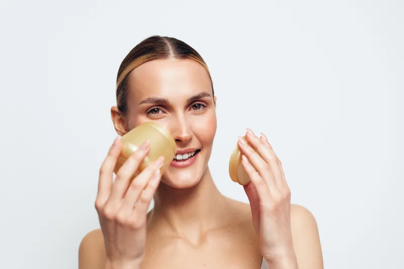 Young woman with clear skin holding two round soap bars, smiling gently, natural beauty concept, clean background, skincare routine, fresh and healthy appearance