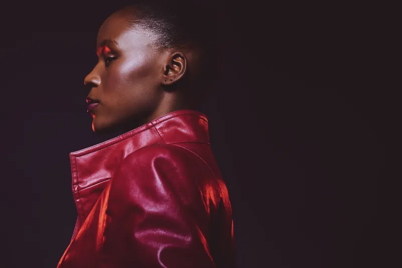 Profile of a young black woman with dark skin wearing a red leather jacket against a dark background. The image highlights fashion, beauty, and confident female portrait concepts.