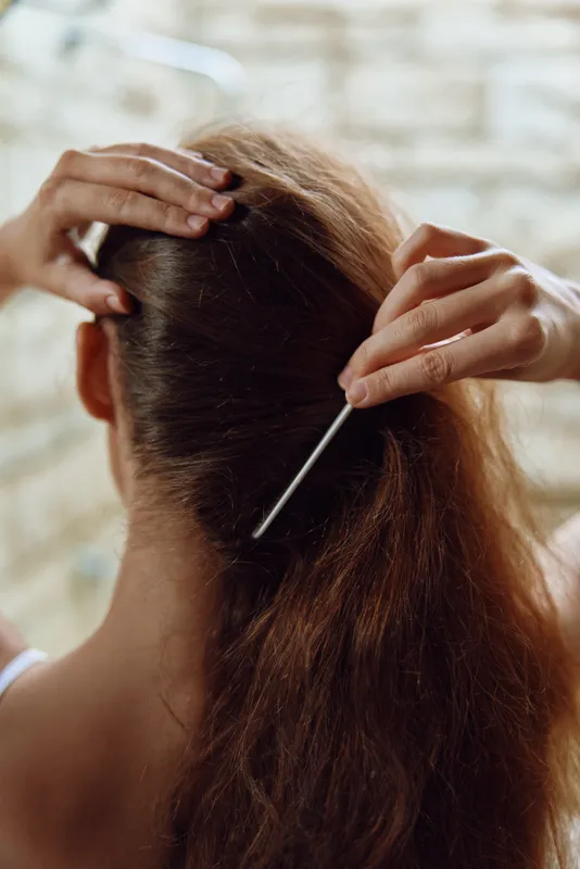woman fixing hair with metal hairpin, close-up view, brown hair, soft natural lighting, indoor background, calm atmosphere