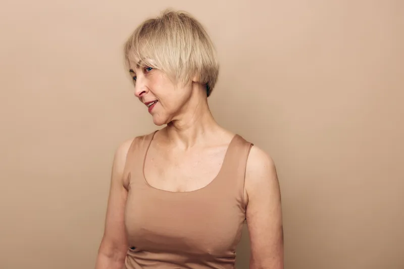 Profile of a mature woman with short blonde hair wearing a beige sleeveless top. Soft neutral background highlights natural beauty, confidence, and aging gracefully concepts.