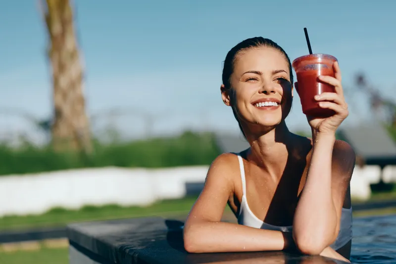Cheerful woman outdoors, smiling by a sunny pool, holding a smoothie drink, casual summer attire, relaxed leisure mood, natural colors, vibrant lifestyle moment