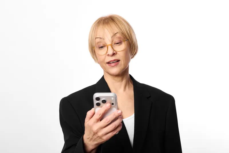 Senior woman with glasses in a black business jacket holds a smartphone, communication on the screen, calm focused mood, isolated portrait on white background, technology