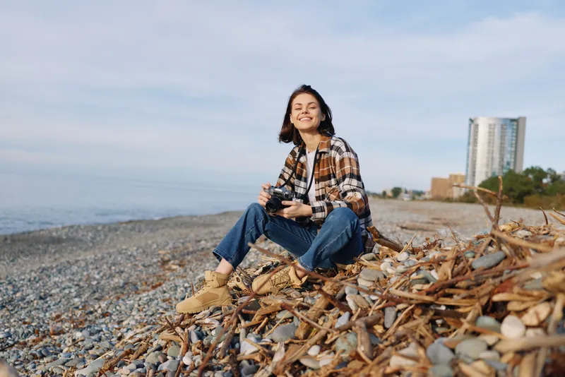 Casual woman photographer on a rocky beach with a camera, smiling under a clear sky, capturing a seaside moment. Dressed in plaid shirt and jeans, with pebbles, water and breezy coastal scenery,