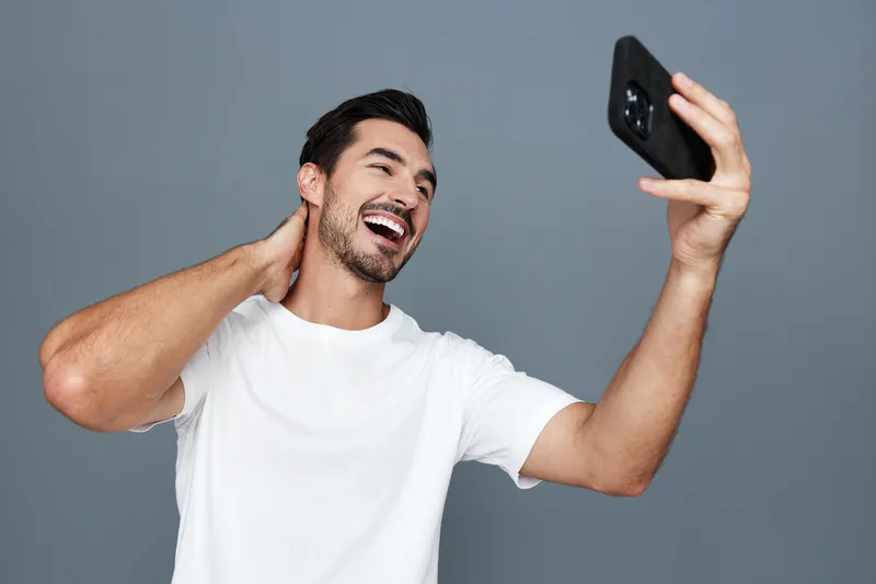 Full body smiling young man taking a selfie with smartphone, wearing white t-shirt, looking joyful and relaxed against a solid gray background. People lifestyle concept.