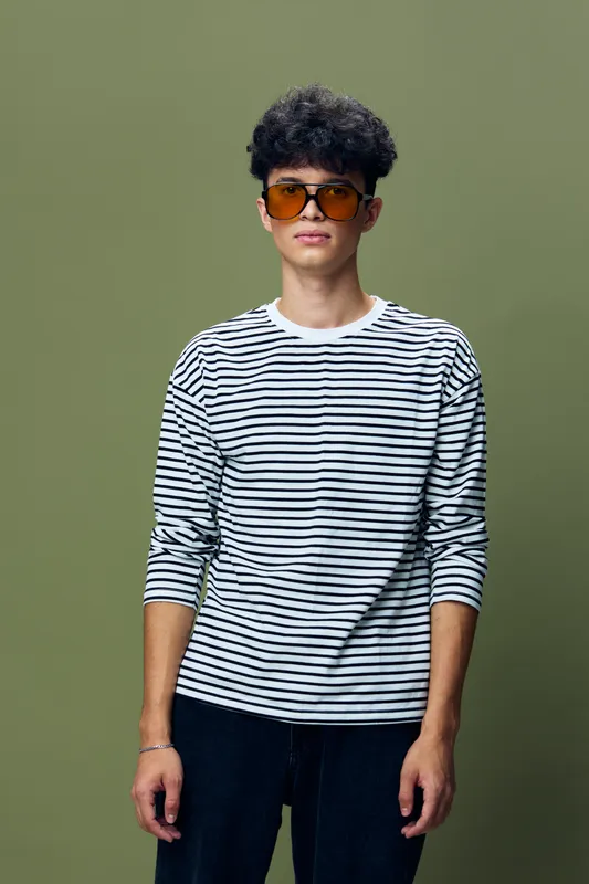 young man wearing striped long sleeve shirt and dark pants stands against olive green background with orange sunglasses, casual style, relaxed pose, modern fashion