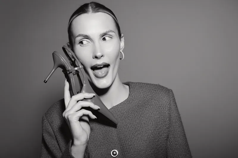 Young Caucasian woman with fair skin holds high heel shoe near her face, looking to the side. Fashion portrait in black and white studio with playful expression and casual outfit.