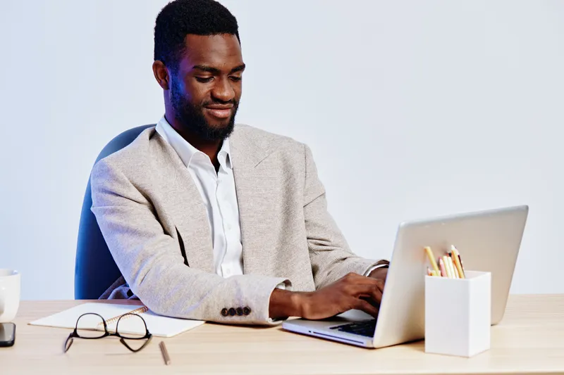 Professional smiling African American man in beige blazer working on a laptop at desk in modern office setting. Business, technology, workplace, focus, adult, casual style, indoors, professional appea