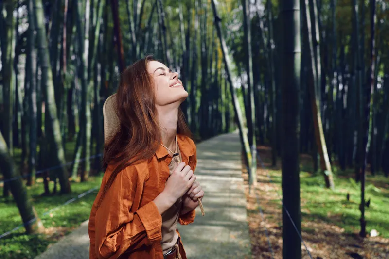 Woman forest path nature happiness portrait outdoors smile enjoying moment with eyes closed and relaxed expression on sunlit trail among tall trees, casual style and peaceful mood