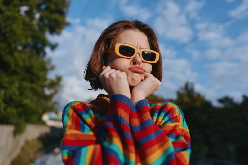 Teenage girl in striped sweater and yellow sunglasses makes a playful pouty face, short brown hair framing an outdoor portrait with suburban background and colorful outfit vibe.