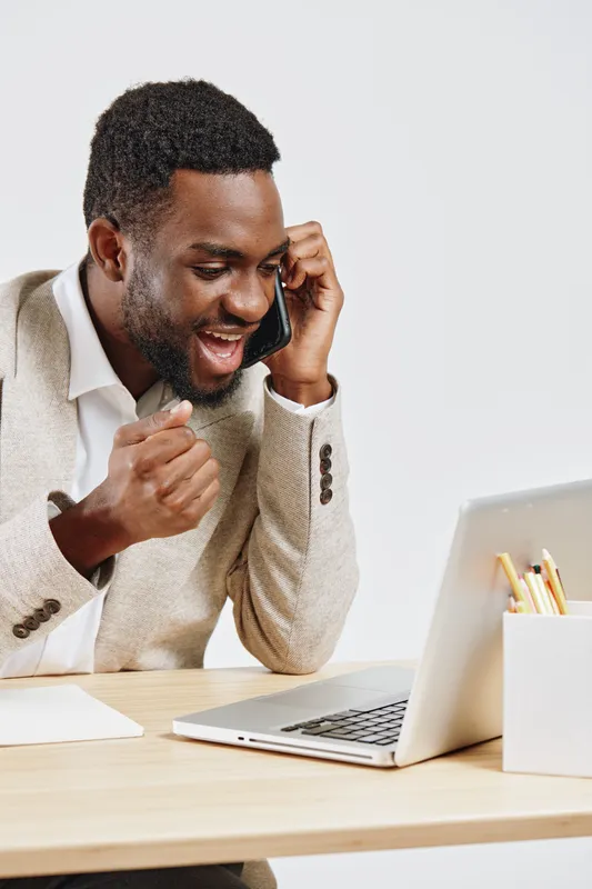 Friendly young African American man in beige blazer using smartphone at work desktop, smiling and looking engaged, indoors with neutral background, professional tone, modern style.