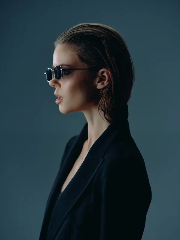 Profile of a stylish woman wearing sunglasses and a black blazer against a dark blue background, expressing confidence and elegance.