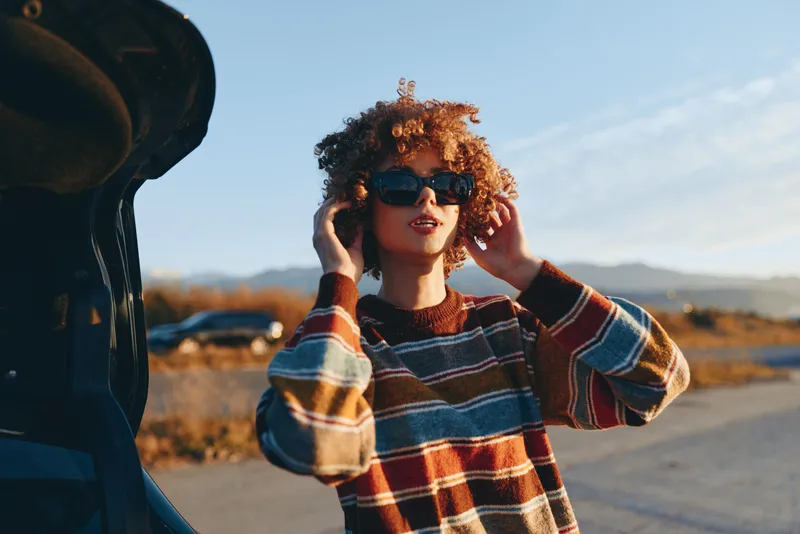 Woman lifestyle outdoor portrait, curly hair, plaid sweater, sunny road background, standing and smiling with hands on head, warm daylight and open landscape, summer travel mood