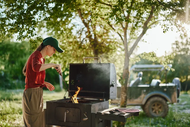 Young woman grilling outdoors in a picturesque setting, wearing casual attire and a cap, showcasing joy and focus while cooking on a sunny day