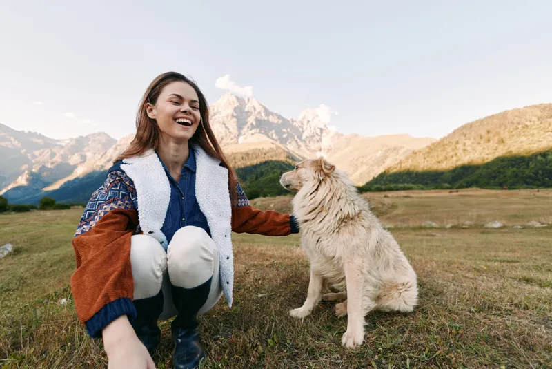 Woman and dog in mountains meadow outdoors, smiling and happy as they play and sit on grass, enjoying pet companionship during a casual hike with natural landscape and soft sunlight.