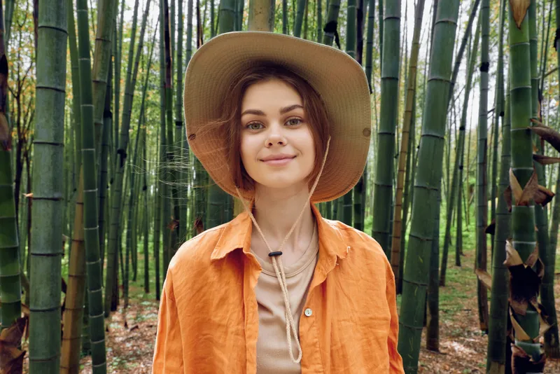 Woman portrait in a bamboo forest wearing a wide hat and orange shirt, smile and calm expression in nature outdoors, soft sunlight, casual travel mood and serene greenery.