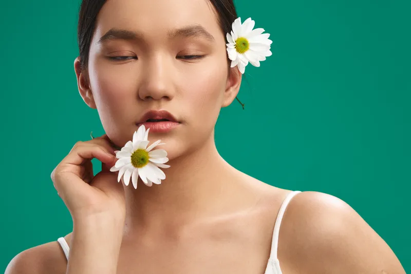 Asian woman with white flowers tucked behind her ear stands before a teal backdrop, exuding serenity and gentle beauty in a minimal, stylish portrait.