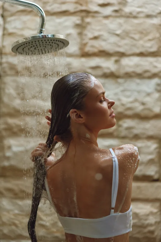 woman showering under rainfall showerhead, washing long wet hair with water splashing, indoors bathroom setting with warm stone wall background