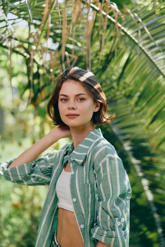 Portrait of a young woman outdoors among tropical foliage, wearing a green striped shirt over a white crop top, softly framed by lush leaves.