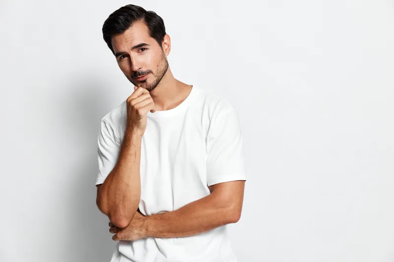 Thoughtful man in a casual white t-shirt poses with a hand on chin, looking at the camera. Studio portrait against a plain light grey background.