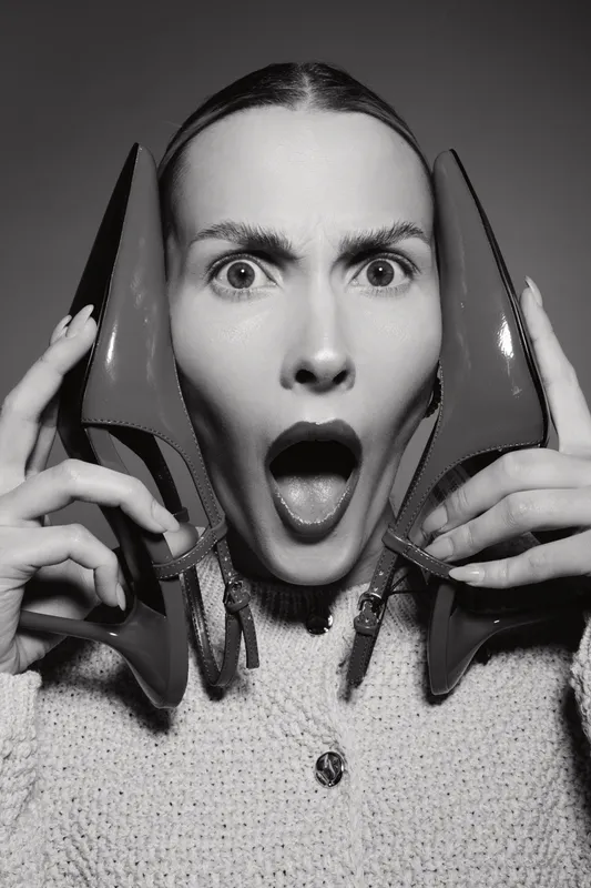 Surprised white woman with fair skin holds red high heel shoes near face, mouth open. Black and white portrait with expressive emotion and fashion concept in studio lighting.