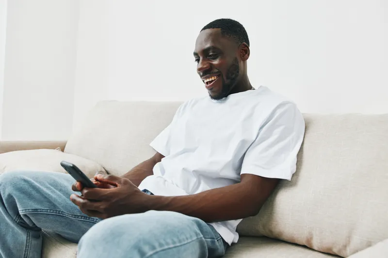 Happy Black man in a white t shirt using a smartphone, sitting on a beige couch, conveying joy and relaxation in a bright, minimalist living space